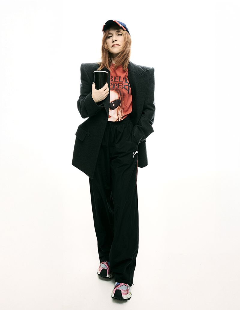 Adult female with long red hair wearing a baseball cap and blazer, holding a black container, stands in a white photo studio.