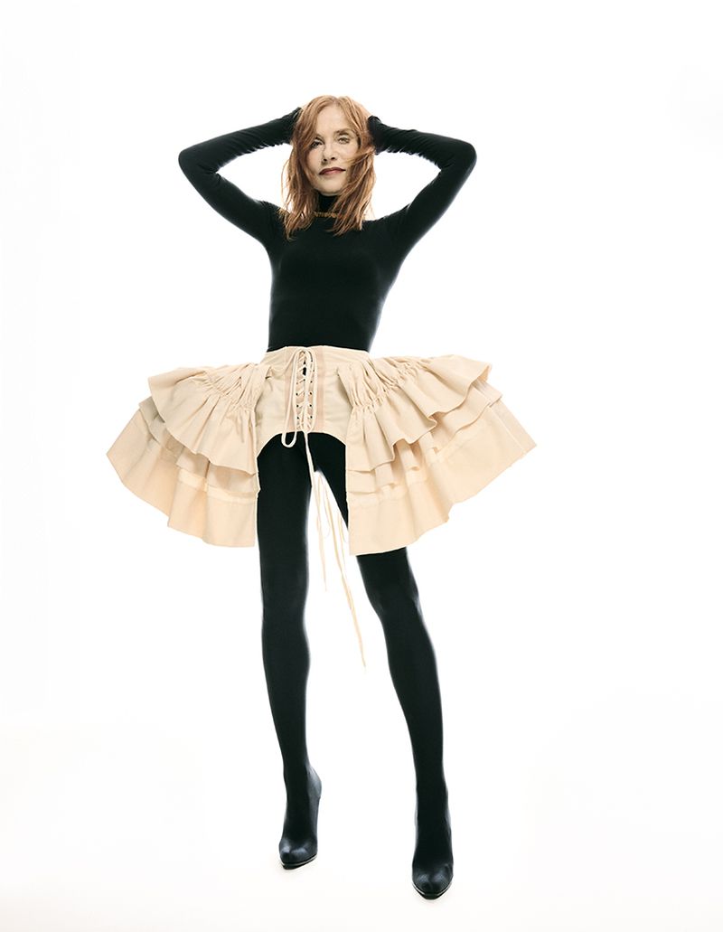 Full-length shot of a woman in a black turtleneck, black leggings, black heels, and a layered ruffled skirt against a white backdrop.