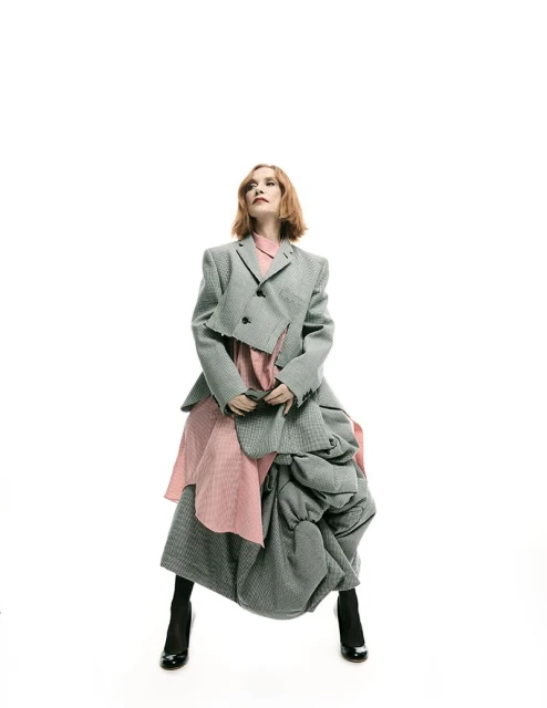 Woman poses in a deconstructed grey jacket and pink dress against a white background, accessorized with black boots, looking upward.