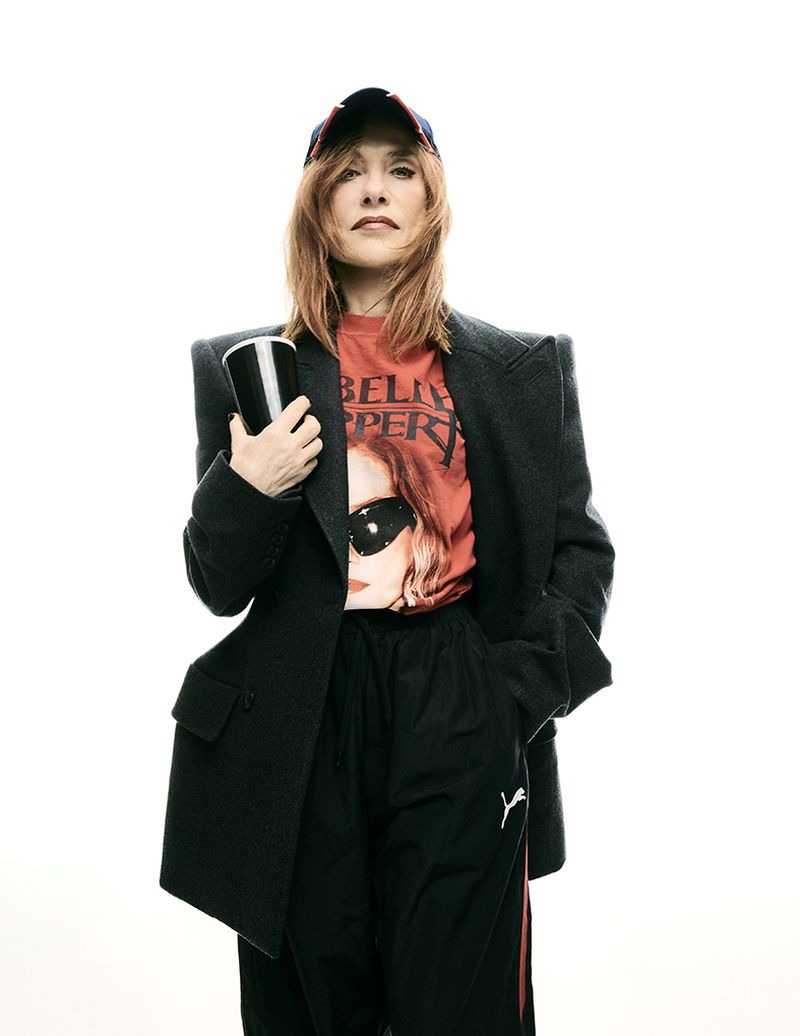 Adult woman in an oversized blazer and baseball cap holds a cup in a studio, posing in front of a white background.