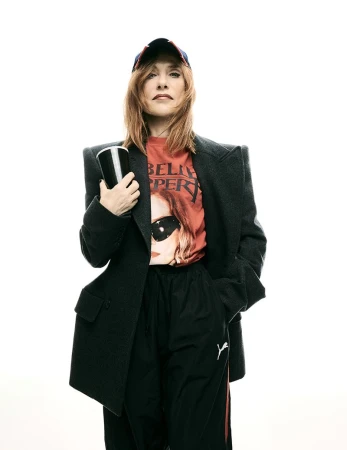 Adult woman in an oversized blazer and baseball cap holds a cup in a studio, posing in front of a white background.