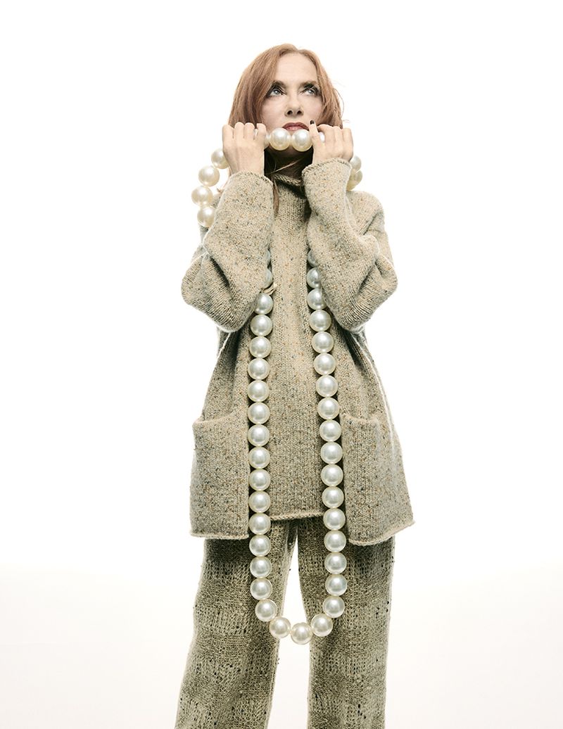 Adult woman in tan knitwear holds a strand of large pearls with a neutral expression against a bright white background in a portrait.
