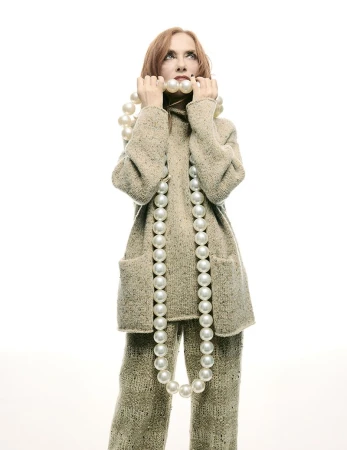 Adult woman in tan knitwear holds a strand of large pearls with a neutral expression against a bright white background in a portrait.