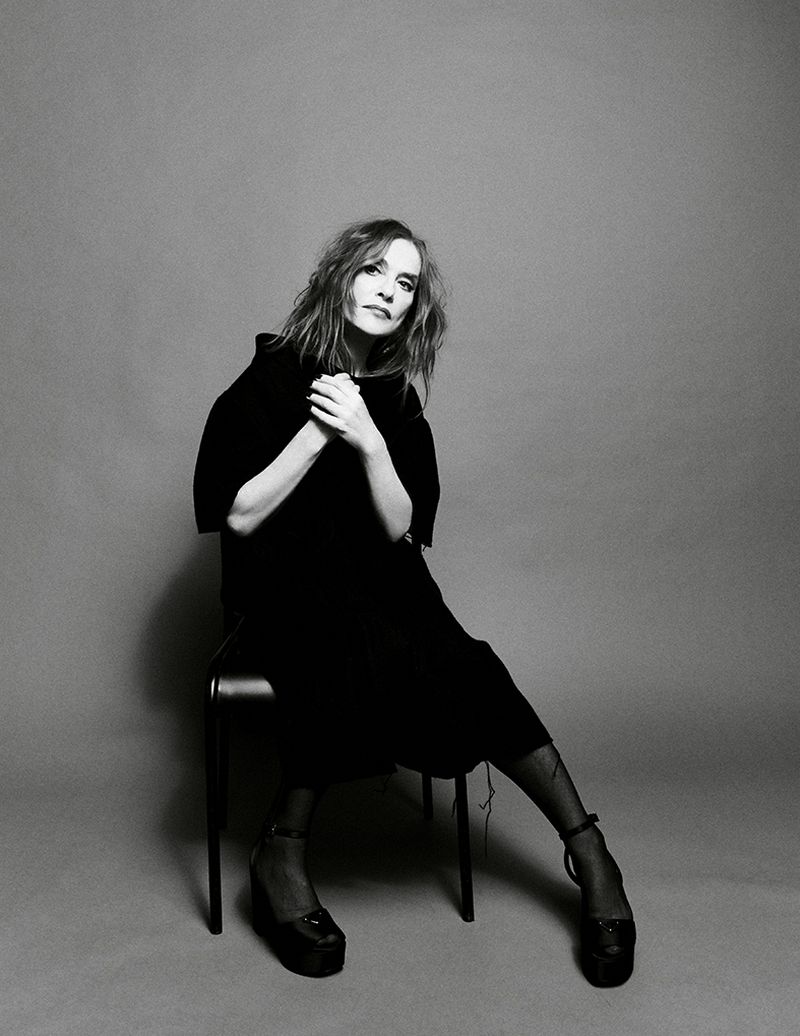 Black and white portrait of a seated adult female in a dark dress and platform shoes against a plain gray background in a photo studio.