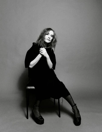 Black and white portrait of a seated adult female in a dark dress and platform shoes against a plain gray background in a photo studio.