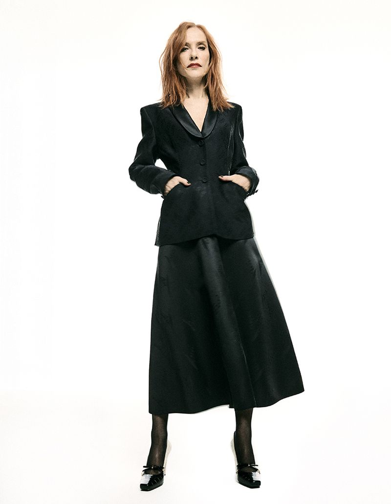 Woman with red hair wearing a black jacket and long skirt against a plain white background