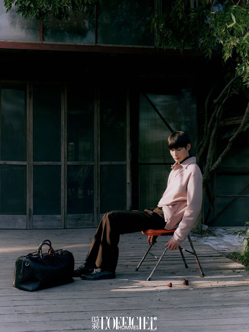 A young adult man sits on a stool with a travel bag on a wooden deck next to an industrial building with climbing foliage.