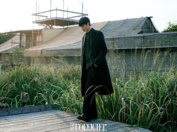 Huang Shixin walks on a wooden platform wearing a long dark tailored coat and trousers surrounded by tall wild grasses.