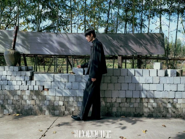 Asian man posing outdoors in black suit next to block wall with bamboo background in editorial fashion shot for L'Officiel Hommes magazine.