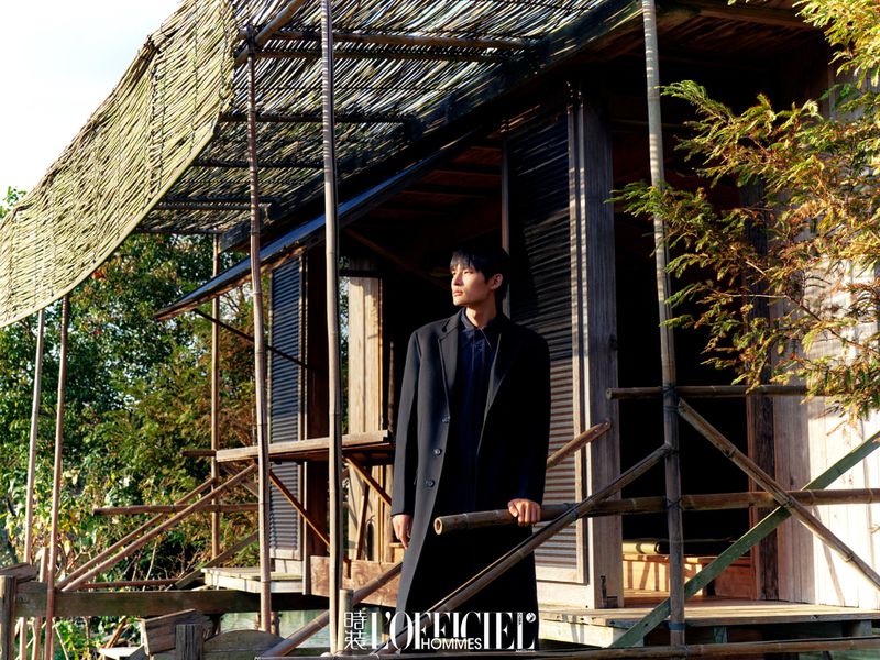 Asian male in black coat poses on rustic wooden porch with natural light. Architecture features weathered design.