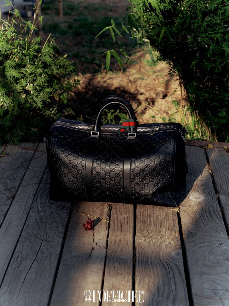 Gucci black leather duffel bag with an embossed monogram pattern on weathered wood planks, placed near green foliage.