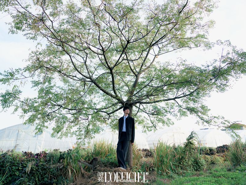 A man stands under a large tree in a field wearing a dark suit, photographed at eye-level on an overcast day for a fashion editorial.