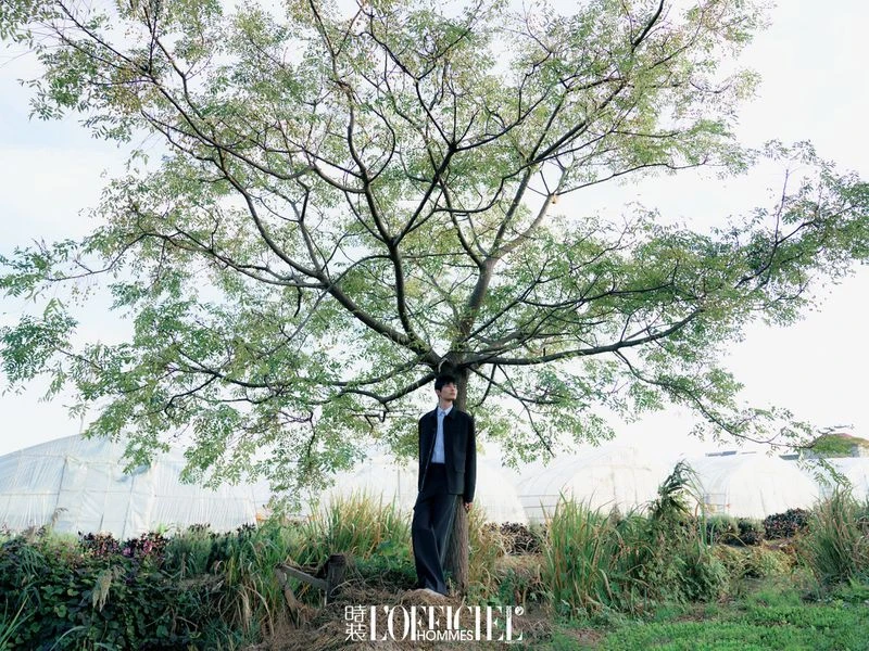 A man stands under a large tree in a field wearing a dark suit, photographed at eye-level on an overcast day for a fashion editorial.