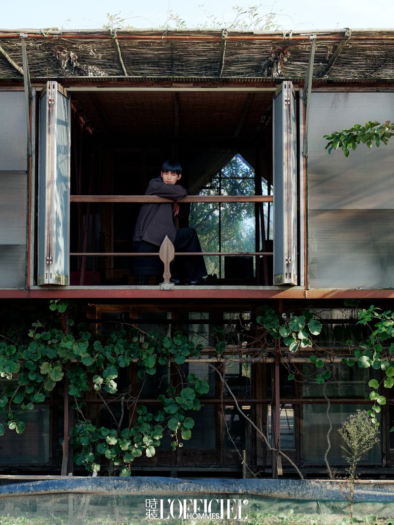 Young man poses at window of rustic two-story structure with climbing plants, shot in natural light for L'Officiel Hommes magazine.