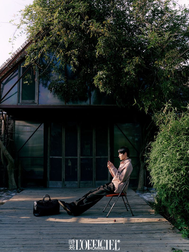 Adult male in a blush jacket sits on a red chair on a wooden deck near a dark building with large trees in an outdoor setting.