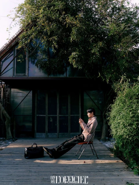 Adult male in a blush jacket sits on a red chair on a wooden deck near a dark building with large trees in an outdoor setting.
