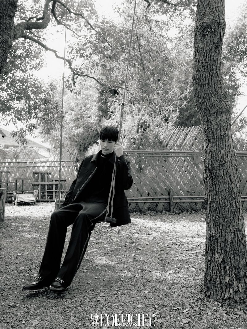 Monochrome portrait of a young man wearing dark clothing, sitting on a rope swing suspended between trees in a rural outdoor setting.