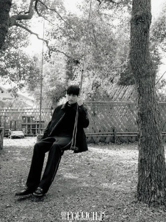 Monochrome portrait of a young man wearing dark clothing, sitting on a rope swing suspended between trees in a rural outdoor setting.