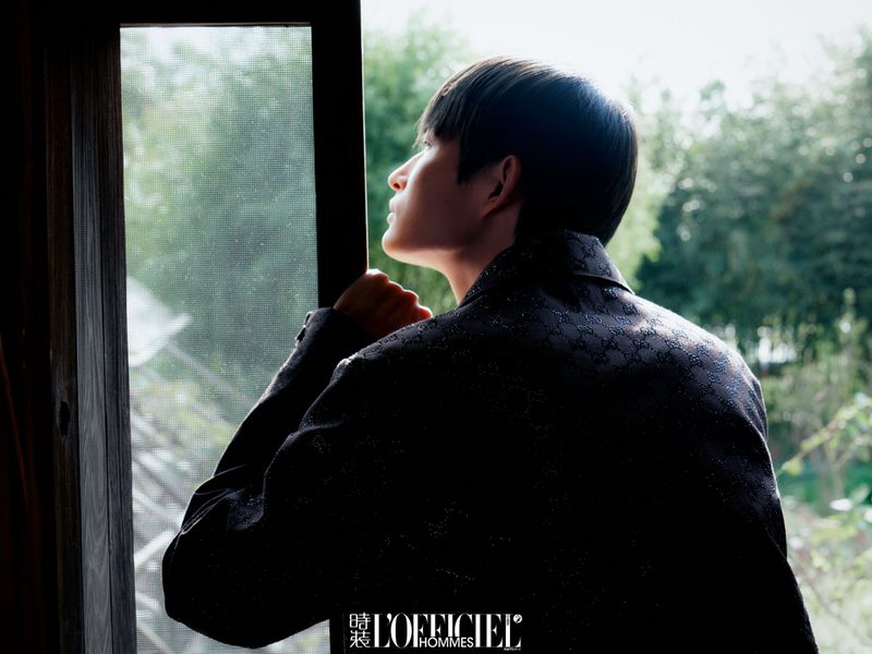 East Asian man in a dark patterned jacket looking out a screened doorway toward lush green trees.