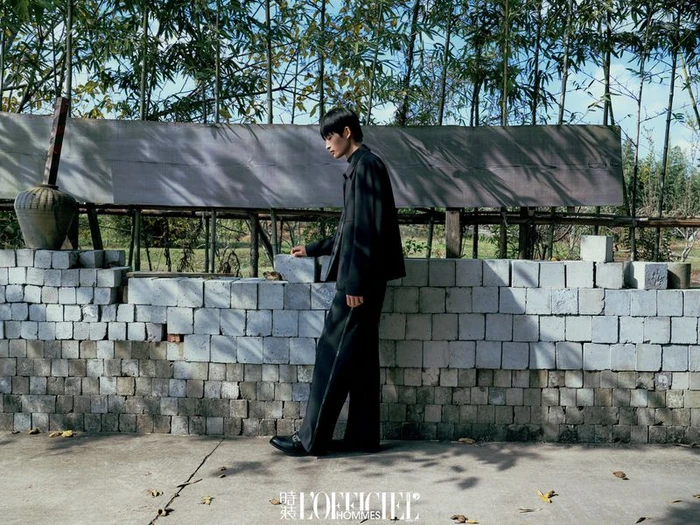 Model wearing dark jacket and trousers walking past a cinder block wall and bamboo grove.