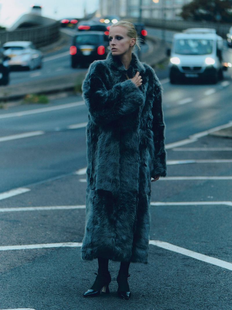 Woman in long fur coat standing in the middle of a city road with traffic, looking left. Dramatic, overcast light and cool color palette.