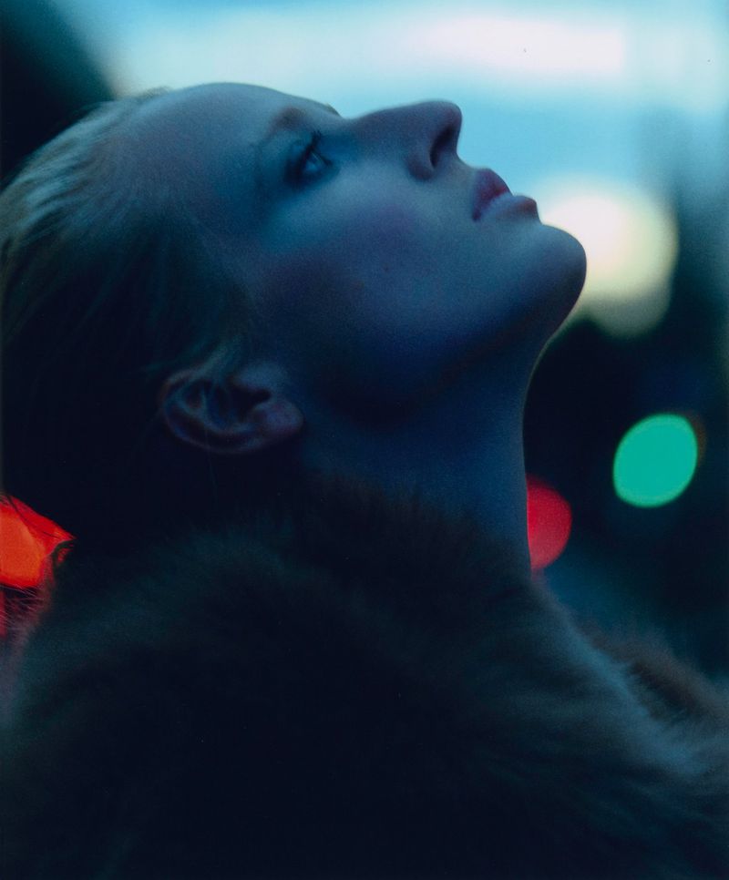 Close-up portrait of a fair-skinned woman looking upwards with a serene expression, set against a blurred background of red and green lights.