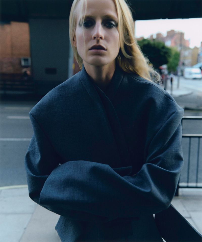 Fashion editorial shot: Woman in a blazer poses outdoors against urban backdrop. Neutral color palette, strong lines, and diffused lighting.