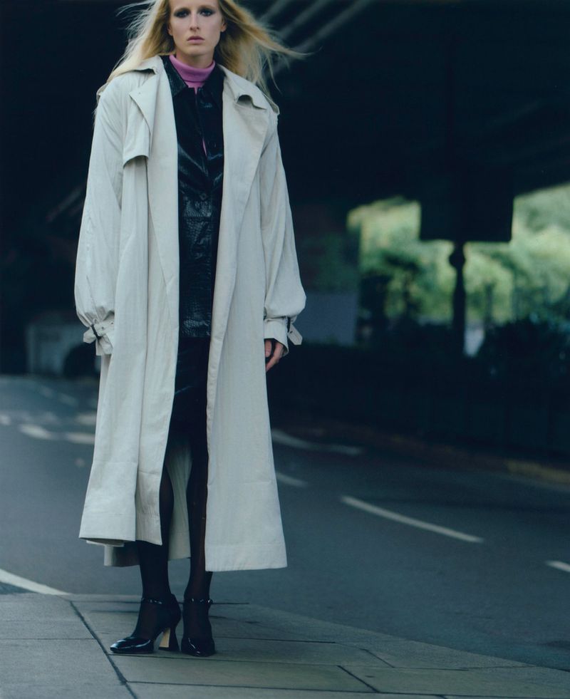A blonde fashion model in a beige trench coat poses on an urban street under an overpass, showcasing a modern and stylish aesthetic.