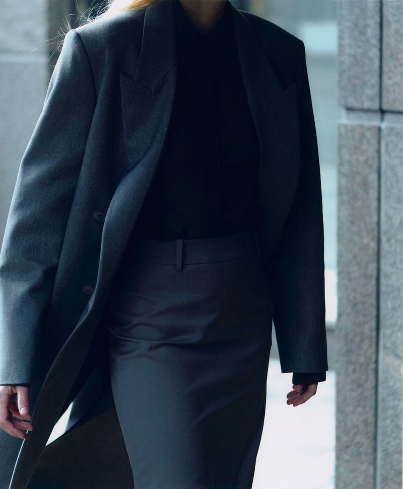 Woman in a grey coat and dark skirt walking in an urban setting with granite walls. Fashionable outerwear and urban style.