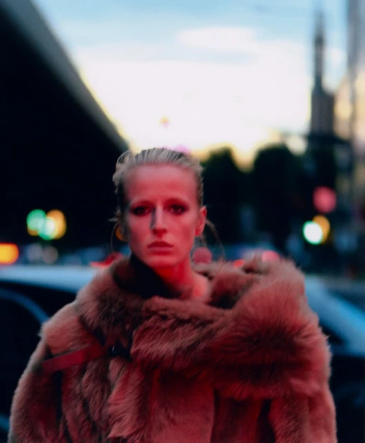 Fashion portrait of a blonde model in a fur coat in an urban setting with blurred background lights, showcasing editorial style.