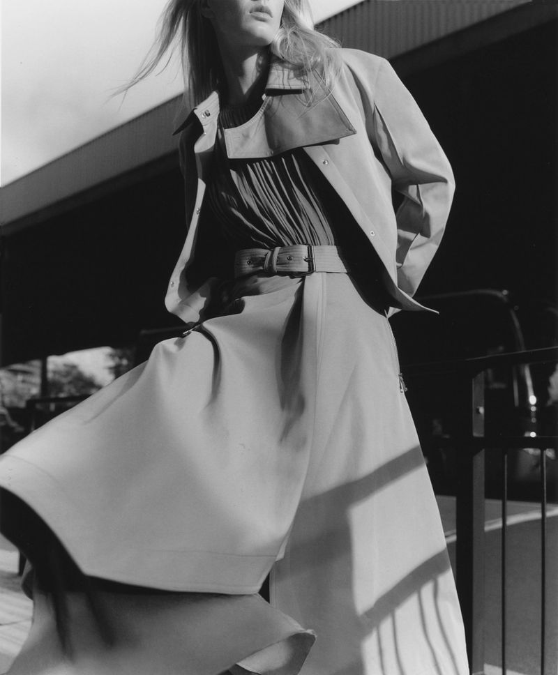 Monochrome outdoor fashion shot of a blond adult woman wearing a belted trench coat and dress with a building in the background.