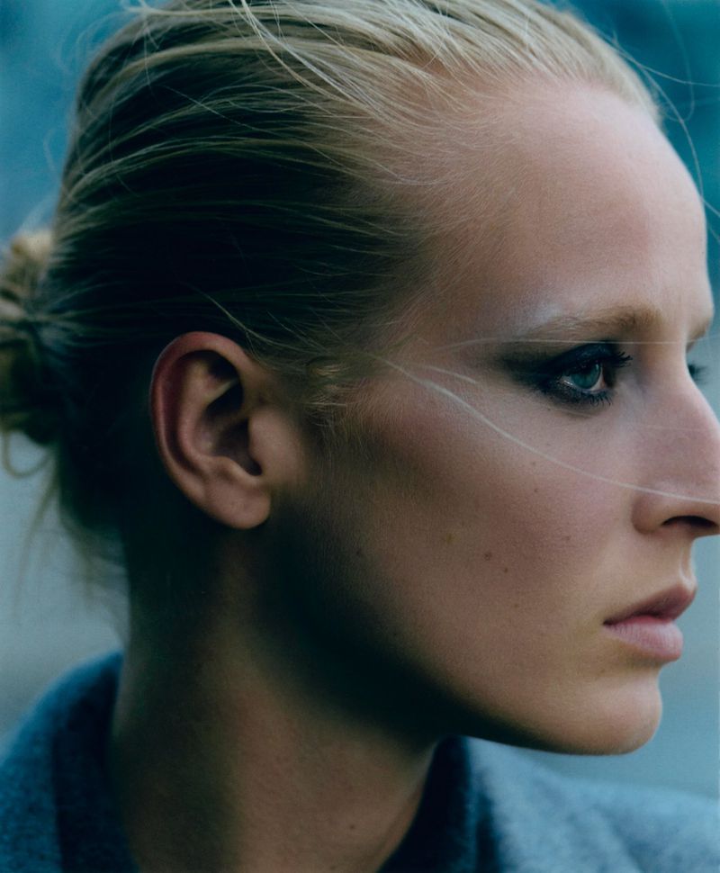 Close-up studio portrait of a fair-skinned adult female with blonde hair, in profile, wearing a grey jacket against a blue backdrop.
