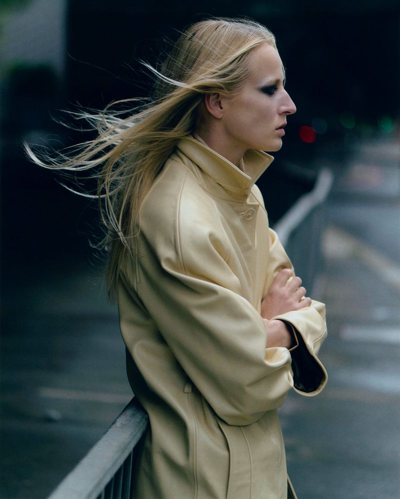 An adult woman with long blonde hair poses outdoors in a light-yellow coat, with her arms crossed and a blurred urban background.