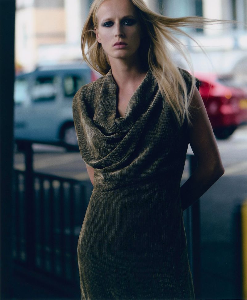 Elegant woman in a shimmering bronze dress poses on a city street, highlighted by natural light, against a backdrop of blurred city traffic.