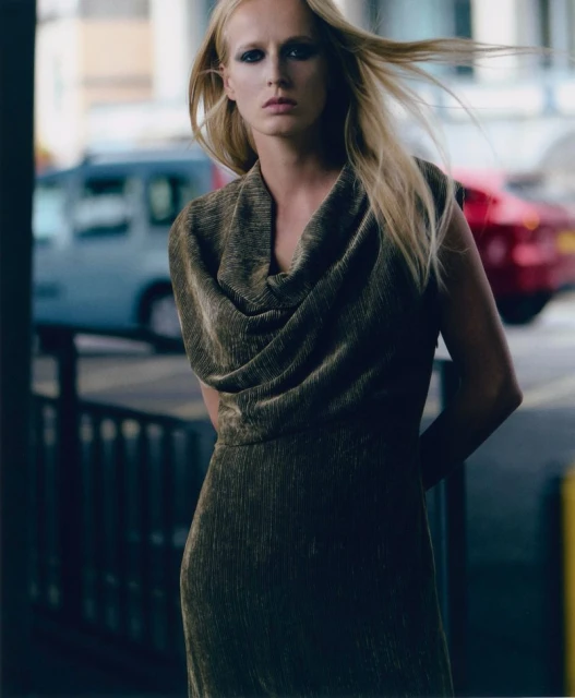 Elegant woman in a shimmering bronze dress poses on a city street, highlighted by natural light, against a backdrop of blurred city traffic.