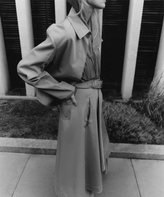 Monochrome fashion shot of a tall, fair-skinned woman in a tan trench coat and skirt, posing outdoors with neutral concrete background.