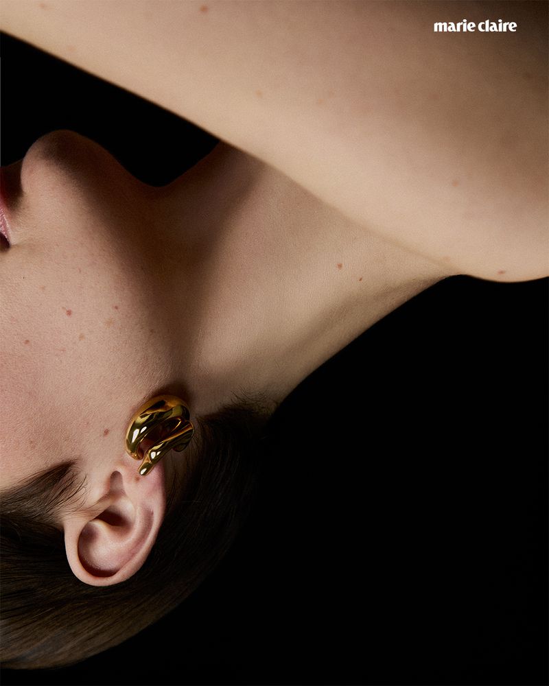 Close-up studio shot of a fair-skinned woman wearing a gold earring with a solid black background; features 'Marie Claire' magazine logo.