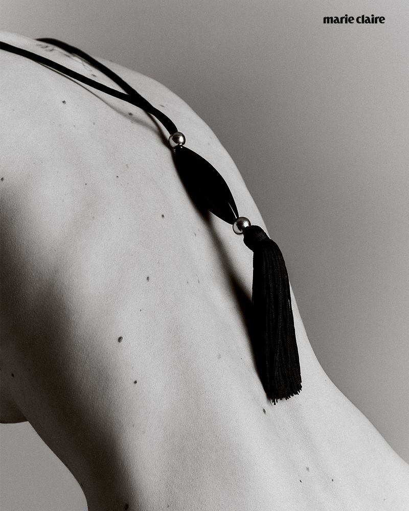 Monochrome shot of a person's back and shoulder adorned with a black tassel necklace and the Marie Claire logo in the upper corner.