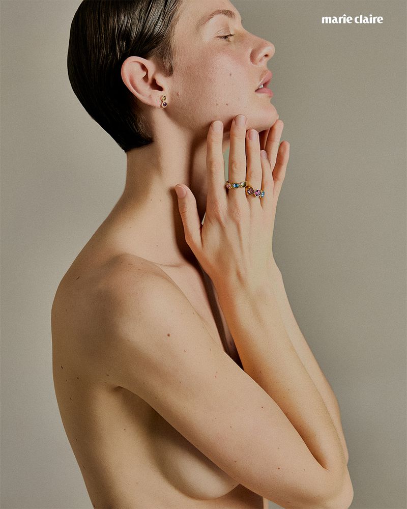 Woman with short dark hair poses with hands on her neck, wearing jewelery in a studio shot with 'Marie Claire' logo.