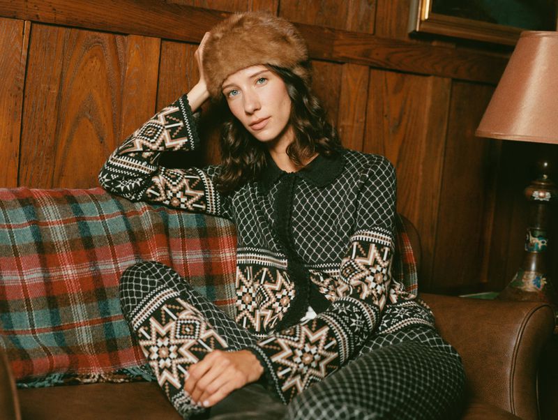 Woman in patterned cardigan and fur hat poses on a brown leather couch in a wood-paneled room with warm lighting.