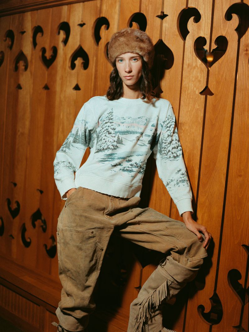 A person in a fur hat and patterned sweater poses against a decorative wooden wall, creating a vintage fashion portrait.