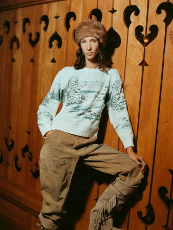A person in a fur hat and patterned sweater poses against a decorative wooden wall, creating a vintage fashion portrait.