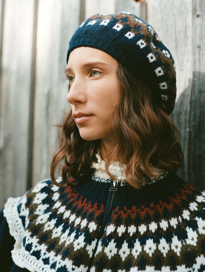 Profile shot of a young woman with wavy brown hair wearing a patterned knit sweater and hat outdoors against a wood background.