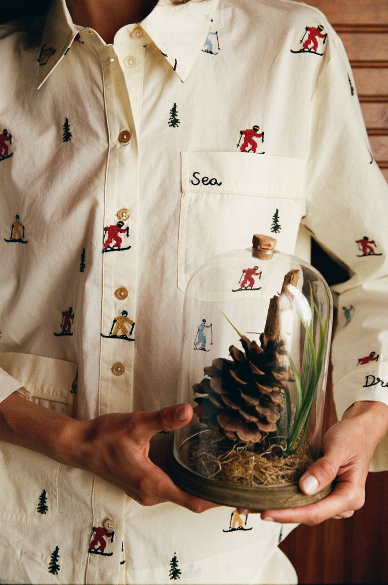 Person holds a pinecone under a glass dome, wearing an embroidered shirt with skier and tree motifs, against a wood background.