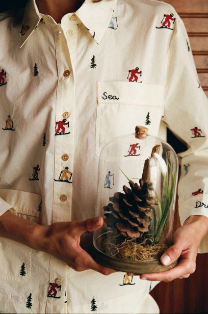 Person holds a pinecone under a glass dome, wearing an embroidered shirt with skier and tree motifs, against a wood background.