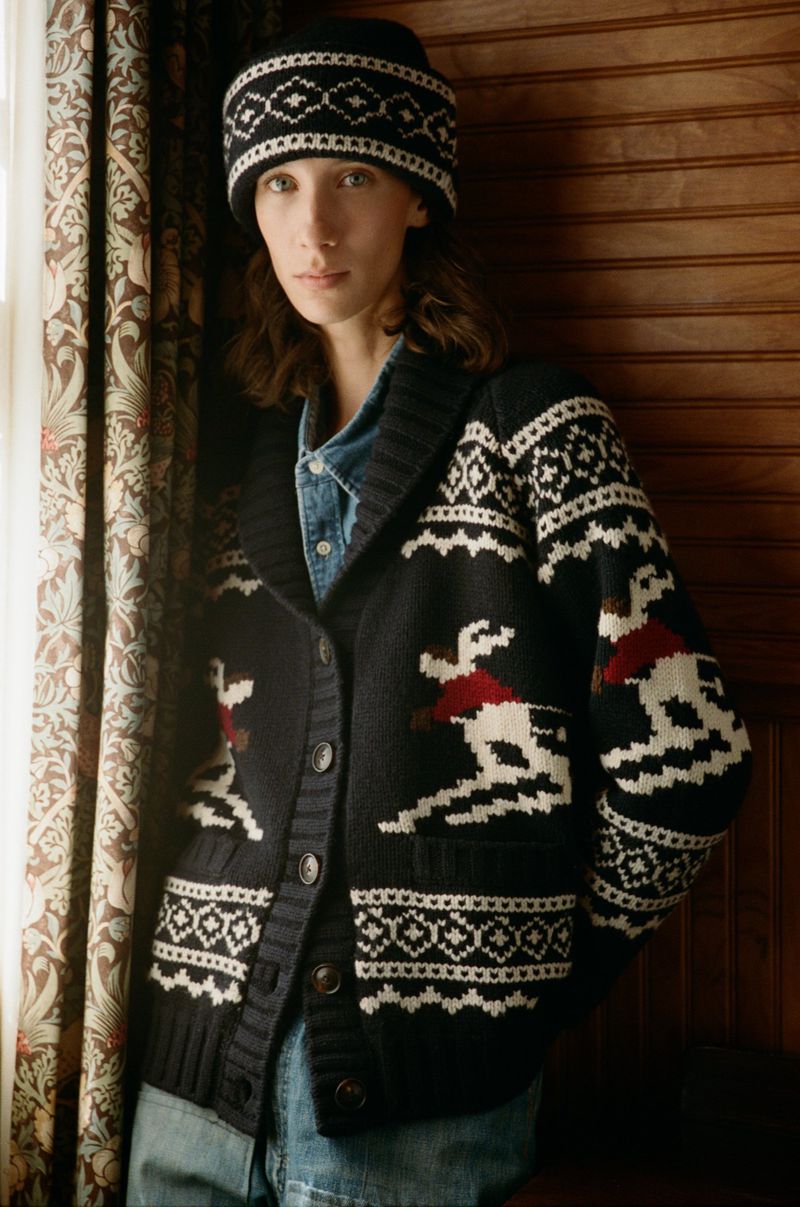 Portrait of a woman wearing a patterned knit hat and cardigan sweater with skier designs, with a vintage aesthetic in an indoor setting.