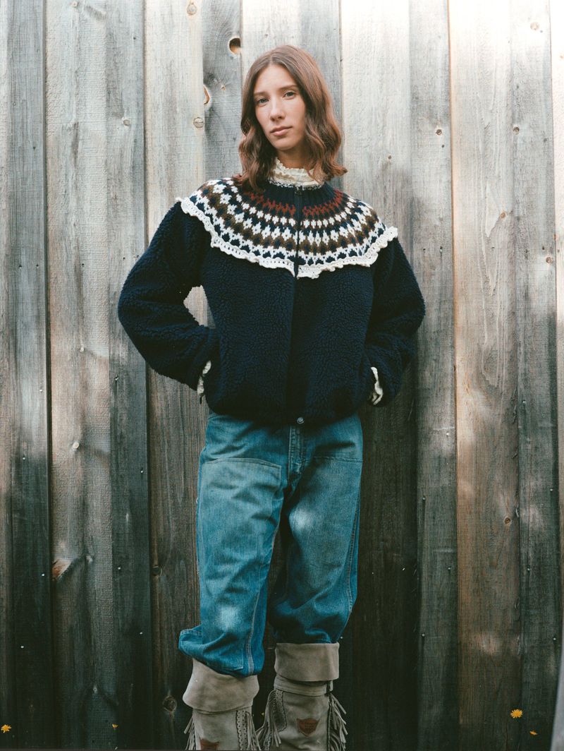 A woman poses in a patterned sweater, jeans, and moccasin boots, standing against a weathered wooden fence in a natural outdoor setting.