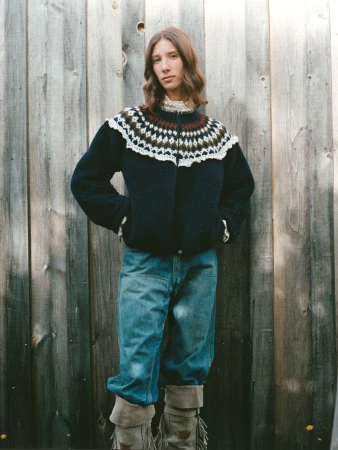 A woman poses in a patterned sweater, jeans, and moccasin boots, standing against a weathered wooden fence in a natural outdoor setting.