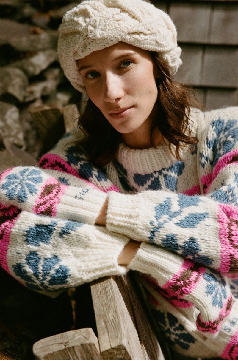 Woman poses outdoors wearing an off-white cable-knit hat and patterned sweater, seated in front of a stone wall and wood.