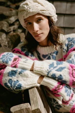 Woman poses outdoors wearing an off-white cable-knit hat and patterned sweater, seated in front of a stone wall and wood.